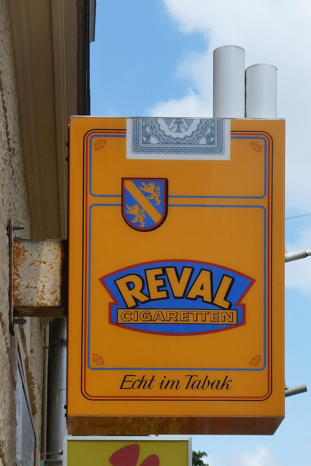 Übergroße Nachbildung der Reval-Packung (Frontseite mit Wappen, Banderole und zwei Zigaretten) als Werbeschild an einem Tabakgeschäft in München, 2023